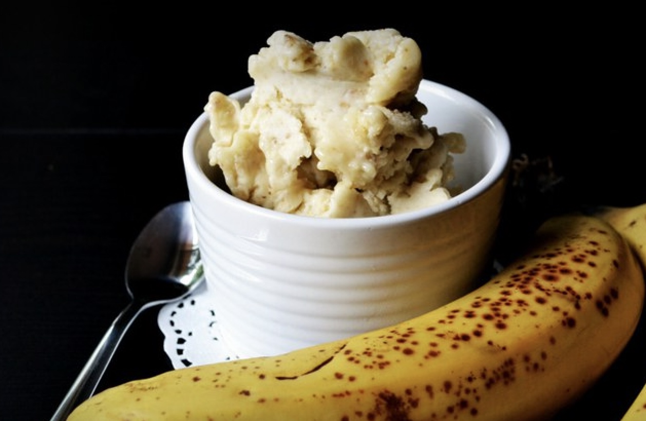 Banana ice cream in a circular white pottery bowl with a black
    background and a banana lying next to it