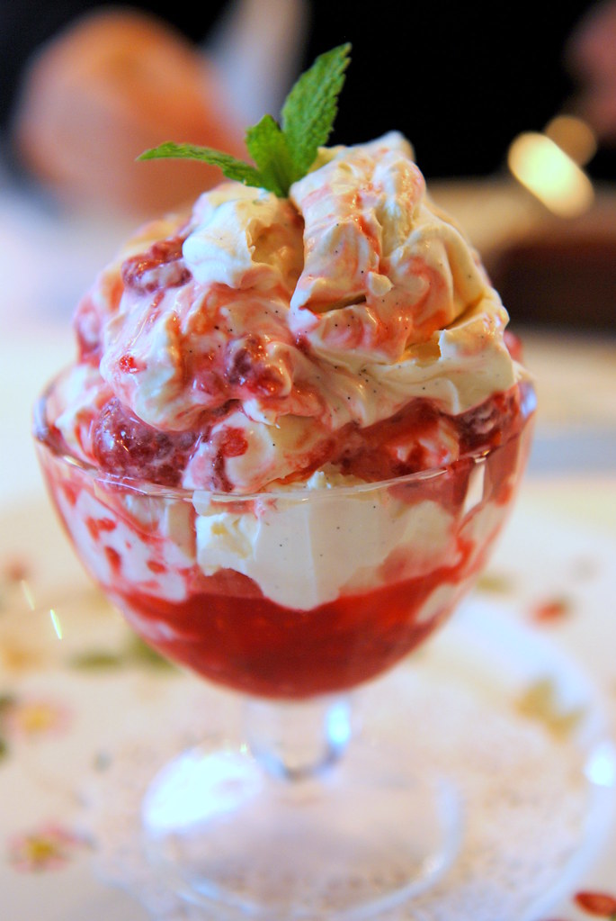 red and white eton mess ice cream in a transparent circular glass bowl with 
    a rounded bottom and a blurred backround of a white tablecloth